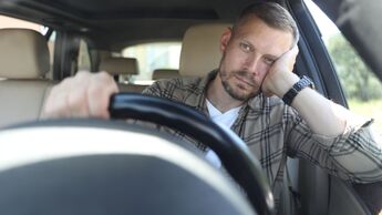 Arbeitnehmer sitzt im Fahrersitz eines Pkw und lehnt den Kopf an die Hand, Blick nach vorn, Innenraum eines Fahrzeugs.