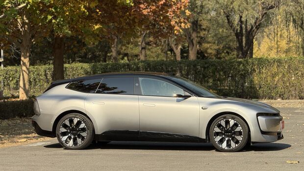 Audi E5 in Grau, Seitenansicht, geparkt im Freien vor Baeumen und Hecken.