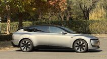 Audi E5 in Grau, Seitenansicht, geparkt im Freien vor Baeumen und Hecken.