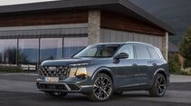 Audi Q7 in blaugrauer Lackierung schräg von vorn vor einem modernen Gebäude mit Berglandschaft.