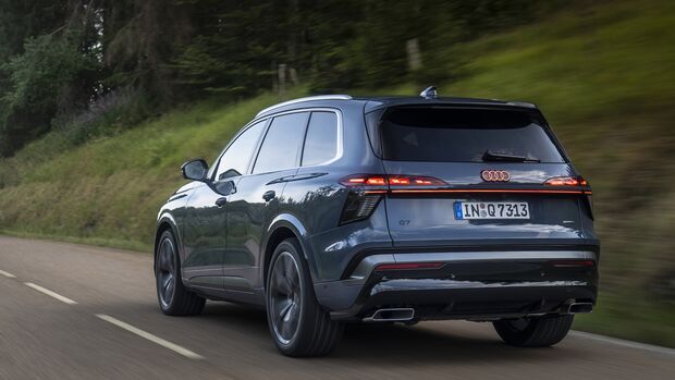 Audi Q7 schräg von hinten während der Fahrt auf einer Landstraße mit Wald und begrüntem Hang im Hintergrund.