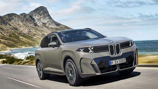 BMW iX xDrive60 in Frontansicht während der Fahrt auf einer Küstenstraße vor Berg- und Meereskulisse.