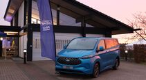 Blauer Ford Transit Custom steht vor einem Gebäude beim Ford Pro Leisure Experience Event in Lommel, neben einer Ford Pro Flagge.