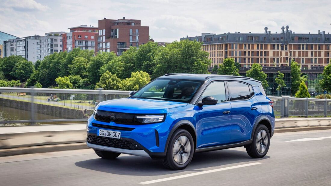 Blauer Opel Frontera SUV fährt auf einer Straße in einer Stadt mit Gebäuden und Grünflächen im Hintergrund.