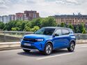 Blauer Opel Frontera SUV fährt auf einer Straße in einer Stadt mit Gebäuden und Grünflächen im Hintergrund.