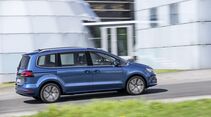 Blauer VW Sharan in Seitenansicht während der Fahrt auf einer Straße vor einem modernen Gebäude.