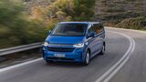 Blauer VW e-Caravelle Elektrovan fährt auf kurviger Landstraße durch hügelige Landschaft bei Tageslicht.