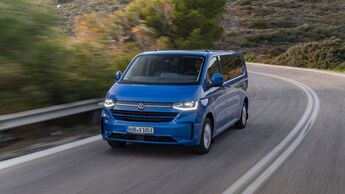 Blauer VW e-Caravelle Elektrovan fährt auf kurviger Landstraße durch hügelige Landschaft bei Tageslicht.