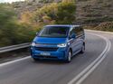 Blauer VW e-Caravelle Elektrovan fährt auf kurviger Landstraße durch hügelige Landschaft bei Tageslicht.