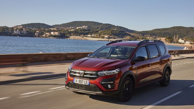 Dacia Jogger in Rot bei der Fahrt auf einer Küstenstraße mit Meer und bewaldeten Hügeln im Hintergrund.