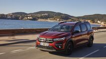 Dacia Jogger in Rot bei der Fahrt auf einer Küstenstraße mit Meer und bewaldeten Hügeln im Hintergrund.
