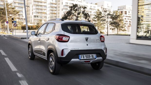 Dacia Spring in Silber, Heckansicht während der Fahrt auf einer städtischen Straße mit Gebäuden im Hintergrund.