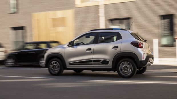 Dacia Spring in Silber, Seitenansicht während der Fahrt auf einer Straße in städtischer Umgebung.