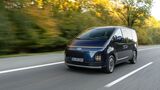 Dunkelblauer Hyundai Staria Elektro in Fahrt aus schräger Frontperspektive mit schmalem Lichtband und vertikalen Scheinwerfern.