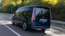 Dunkelblauer Hyundai Staria Elektro während der Fahrt in schräger Heckansicht mit großer Heckscheibe und vertikalen Rückleuchten.