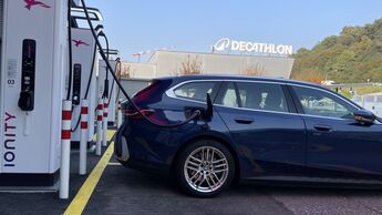 Ein Elektroauto lädt an einer Ionity-Schnellladestation, im Hintergrund ein Decathlon-Markt und herbstliche Umgebung.