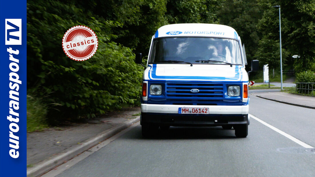 Ein blau-weißer Ford Transit Oldtimer fährt frontal auf einer Straße, umgeben von Bäumen. Links ist das eurotransportTV-Logo zu sehen, daneben ein „Classics“-Stempel.