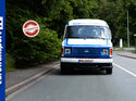 Ein blau-weißer Ford Transit Oldtimer fährt frontal auf einer Straße, umgeben von Bäumen. Links ist das eurotransportTV-Logo zu sehen, daneben ein „Classics“-Stempel.
