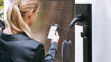 Eine Person bedient eine App vor einer Wallbox zum Laden eines Elektroautos am Wohnhaus.