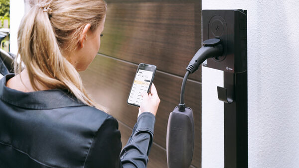 Eine Person bedient eine App vor einer Wallbox zum Laden eines Elektroautos am Wohnhaus.