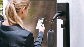 Eine Person bedient eine App vor einer Wallbox zum Laden eines Elektroautos am Wohnhaus.