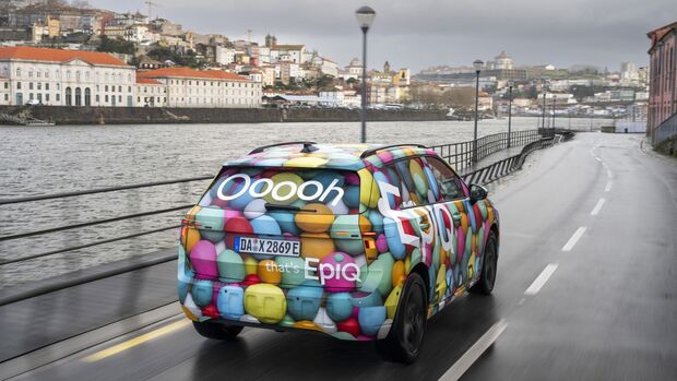 Elektrischer Skoda Epiq mit farbigem Tarnfoliendesign in Heckansicht auf einer Straße entlang eines Flusses.