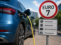 Elektroauto an einer Ladesäule neben einem Euro-7-Schild mit Hinweisen auf Bremsstaub, Reifenabrieb und Fahrzeugdaten.