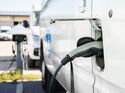 Elektrotransporter lädt an einer Ladestation auf Betriebsgelände, weiteres Nutzfahrzeug im Hintergrund.