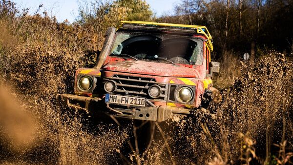 Feuerwehr-INEOS-Grenadier fährt frontal durch tiefen Schlamm und wirbelt große Schmutzmengen auf.
