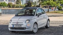 Fiat 500 Electric in Silber auf Pflastersteinfläche am Wasser mit Stadtgebäuden im Hintergrund.