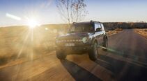 Ford Bronco fährt auf asphaltierter Straße bei tief stehender Sonne, Frontansicht mit eingeschalteten Scheinwerfern.