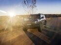 Ford Bronco fährt auf asphaltierter Straße bei tief stehender Sonne, Frontansicht mit eingeschalteten Scheinwerfern.