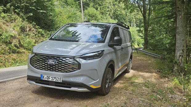 Ford E-Transit Custom AWD Trail in Frontansicht auf einem Waldparkplatz mit markantem Kühlergrill.