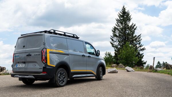 Ford E-Transit Custom AWD Trail in Heckansicht mit Dachträger auf einem Parkplatz in natürlicher Umgebung.