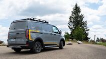 Ford E-Transit Custom AWD Trail in Heckansicht mit Dachträger auf einem Parkplatz in natürlicher Umgebung.