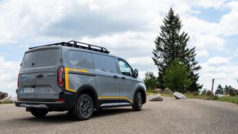 Ford E-Transit Custom AWD Trail in Heckansicht mit Dachträger auf einem Parkplatz in natürlicher Umgebung.