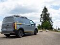 Ford E-Transit Custom AWD Trail in Heckansicht mit Dachträger auf einem Parkplatz in natürlicher Umgebung.