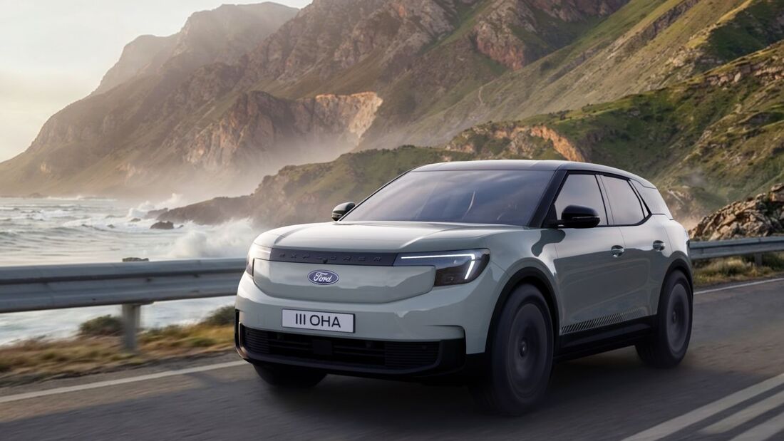 Ford Explorer Elektro-SUV fährt auf Küstenstraße entlang von Meer und Bergen, Frontansicht während der Fahrt.