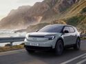 Ford Explorer Elektro-SUV fährt auf Küstenstraße entlang von Meer und Bergen, Frontansicht während der Fahrt.