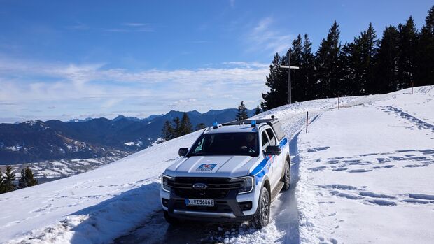 Ford Ranger Plug-in-Hybrid der Bergwacht Bayern auf verschneiter Bergstraße nahe einem Gipfelkreuz.