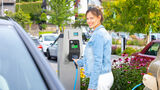 Frau hält Ladekabel an Mennekes AMTRON Wallbox auf einem Parkplatz im Freien neben einem Elektroauto.