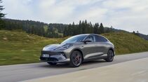 Grauer MG IM6 in Seiten- und Frontansicht während der Fahrt auf einer Straße in einer bewaldeten Hügellandschaft.