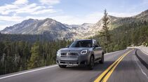 Grauer MINI Countryman Untamed Edition fährt auf einer asphaltierten Bergstraße mit doppelter gelber Mittellinie vor bewaldeten Bergen.