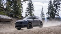 Grauer MINI Countryman Untamed Edition fährt auf einer staubigen Schotterpiste durch einen Nadelwald, aufgenommen schräg von vorn.