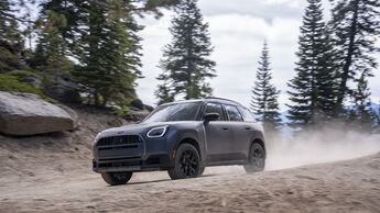 Grauer MINI Countryman Untamed Edition fährt auf einer staubigen Schotterpiste durch einen Nadelwald, aufgenommen schräg von vorn.