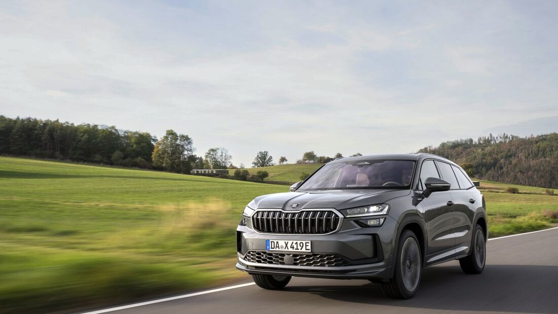 Grauer Skoda Kodiaq SUV in Front-Dreiviertelansicht während der Fahrt auf einer Landstraße mit Wiesen und Bäumen im Hintergrund.