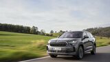 Grauer Skoda Kodiaq SUV in Front-Dreiviertelansicht während der Fahrt auf einer Landstraße mit Wiesen und Bäumen im Hintergrund.