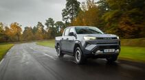 Grauer Toyota Hilux BEV Elektro-Pick-up fährt auf nasser Landstraße durch Waldgebiet.