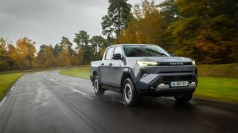 Grauer Toyota Hilux BEV Elektro-Pick-up fährt auf nasser Landstraße durch Waldgebiet.