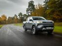 Grauer Toyota Hilux BEV Elektro-Pick-up fährt auf nasser Landstraße durch Waldgebiet.